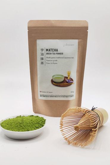 Premium grade Japanese Matcha powder displayed in a dish with a bamboo whisk, next to J-okini’s 100g Matcha package. Pesticide-free and perfect for traditional tea, Matcha lattes, and sweets. Available at j-okini.com in Malta
