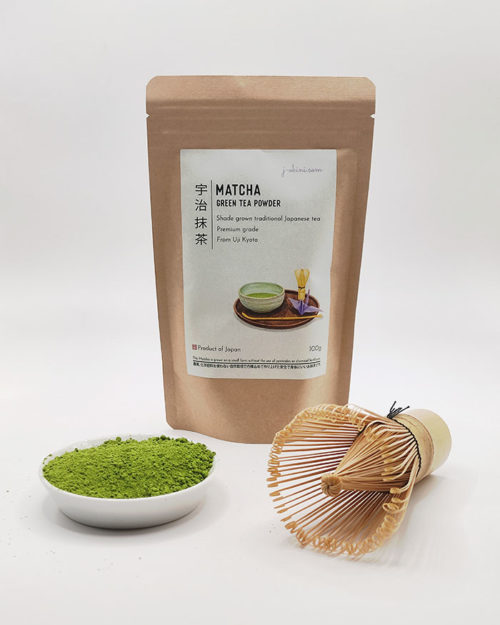 Premium grade Japanese Matcha powder displayed in a dish with a bamboo whisk, next to J-okini’s 100g Matcha package. Pesticide-free and perfect for traditional tea, Matcha lattes, and sweets. Available at j-okini.com in Malta