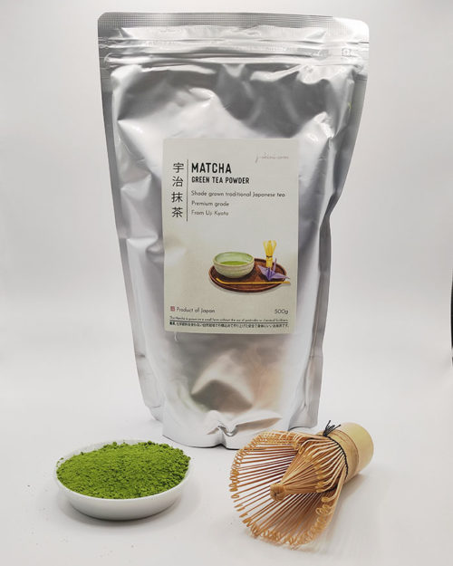 Premium grade Japanese Matcha powder displayed in a dish with a bamboo whisk, in front of J-okini’s 500g Matcha package. Pesticide-free and perfect for traditional tea, Matcha lattes, and sweets. Available at j-okini.com in Malta