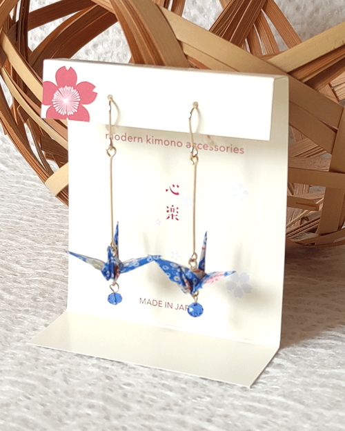 Crafted with meticulous attention to detail, these earrings feature a 3cm piece of Chiyogami, a traditional Japanese washi paper, known for its vibrant and intricate designs. Each pair is carefully sealed with three layers of protective coating to ensure durability and shine. At the heart of these earrings is the motif of the Tsuru, the Japanese word for crane. Revered in Japanese culture, the Tsuru is celebrated as a symbol of peace and longevity, embodying wishes for prosperity and health. These earrings are not only a testament to unique craftsmanship but also carry a deep cultural significance. The design is complemented by a delicate blue glass bead, harmonizing beautifully with the blue Chiyogami paper to create a piece that is both eye-catching and meaningful. Made in Japan. Available at j-okini.com