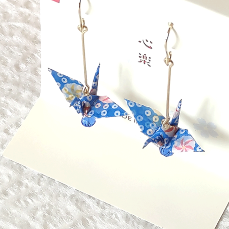 Crafted with meticulous attention to detail, these earrings feature a 3cm piece of Chiyogami, a traditional Japanese washi paper, known for its vibrant and intricate designs. Each pair is carefully sealed with three layers of protective coating to ensure durability and shine. At the heart of these earrings is the motif of the Tsuru, the Japanese word for crane. Revered in Japanese culture, the Tsuru is celebrated as a symbol of peace and longevity, embodying wishes for prosperity and health. These earrings are not only a testament to unique craftsmanship but also carry a deep cultural significance. The design is complemented by a delicate blue glass bead, harmonizing beautifully with the blue Chiyogami paper to create a piece that is both eye-catching and meaningful. Made in Japan. Available at j-okini.com