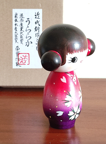This 近代創作こけし modern creative Kokeshi doll is called 'うららか（おだんご）- Uraraka (Odango)' and it was created by an award-winning artist 'ちえ Chie'. The doll's feature would be the space buns.  Her Kimono has a design of Sakura (cherry blossoms) and the gradation of red to purple is beautiful. Find it at j-okini.com in Malta