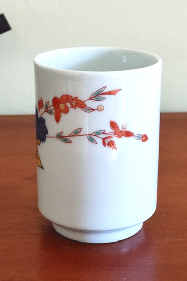 vintage teacup, crafted from white porcelain, features a  delicate hand-painted design of vibrant green, yellow, and blue leaves with a red flower gracefully extending toward the rim.. Available at j-okini.com in Malta