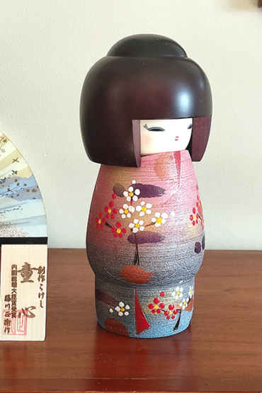Modern Creative Kokeshi doll named Doshin, designed by artist Fujikawa Shoei, standing 20 cm tall and dressed in a pink and mauve kimono with red and white plum blossoms, symbolizing the innocence and purity of a child’s heart.. Buy it at j-okini.com in Malta