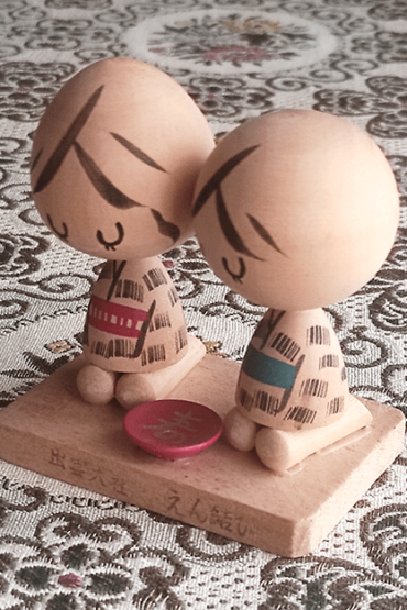 A pair of vintage wooden dolls from Izumo Taisha Shrine in Seiza position, representing a boy and girl. Hand-painted with traditional kimonos and seated with a red sake cup inscribed with "寿" (kotobuki). A faint craftsman's stamp is next to the girl on the wooden base.