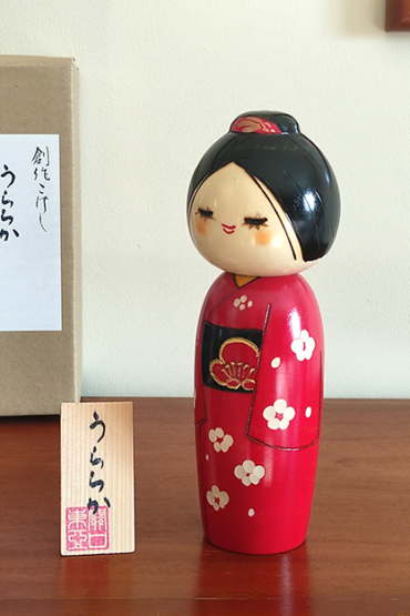 Modern Creative Kokeshi doll named Uraraka, crafted by award-winning artist Sekiguchi Toua, wearing a vibrant red kimono with plum blossom patterns, a black obi with matching design, and a plum blossom Kanzashi hair ornament in a stylish bun.. Buy it online at j-okini.com in Malta