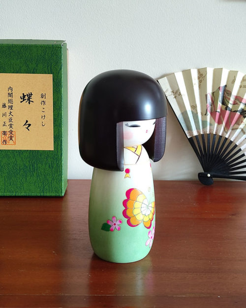 Handcrafted Japanese Kokeshi doll 'Chouchou' by artist Fujikawa Shoei, featuring a light green Michiyuki kimono jacket with a butterfly and pink floral design. Available at j-okini.com in Malta