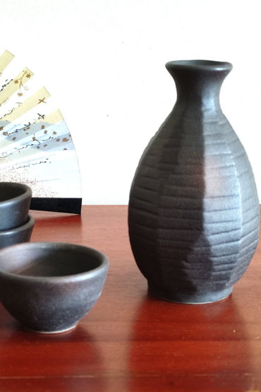 A Sogi Gata Bizen Sake Set featuring a textured black tokkuri bottle with a scraped design and four matching cups, displayed on a wooden surface with a Japanese folding fan in the background. Made in Japan. Available at j-okini.com in Malta