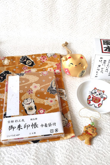 Lucky Cat Blessings Set featuring a golden brown Japanese Goshuincho accordion notebook with a matching drawstring pouch, a white porcelain Maneki-neko small dish, a golden Banko Ware Feng Shui Maneki-neko ceramic figurine, and a handmade Chirimen Cats’ Netsuke strap. A charming collection of Japanese lucky cat-themed items, perfect for gifts or personal good fortune. Made in Japan. Available at j-okini.com in Malta