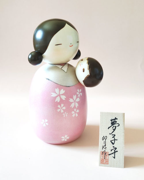 Standing at 19.5 cm tall, this large creative Kokeshi features a mother gently cradling her baby inside a shimmering silver and pink haori. The sparkling pink hue adds a soft, dreamlike glow, while delicate sakura (cherry blossom) motifs adorn the robe. j-okini.com in Malta