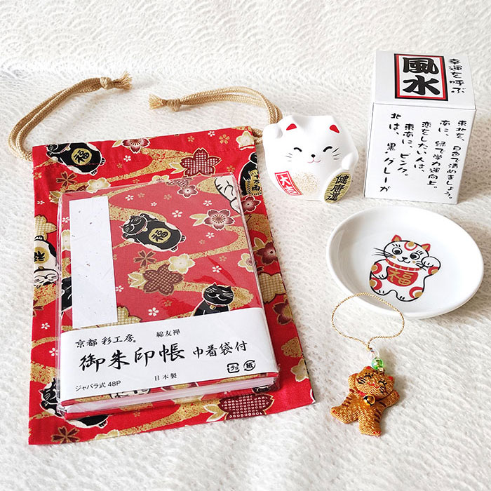 Lucky Cat Blessings Set featuring a red Japanese Goshuincho accordion notebook with a matching drawstring pouch, a white porcelain Maneki-neko small dish, a Banko Ware Feng Shui white Maneki-neko ceramic figurine, and a handmade Chirimen Cats’ Netsuke strap. A charming collection of Japanese lucky cat-themed items, perfect for gifts or personal good fortune. Made in Japan. Available at j-okini.com in Malta.