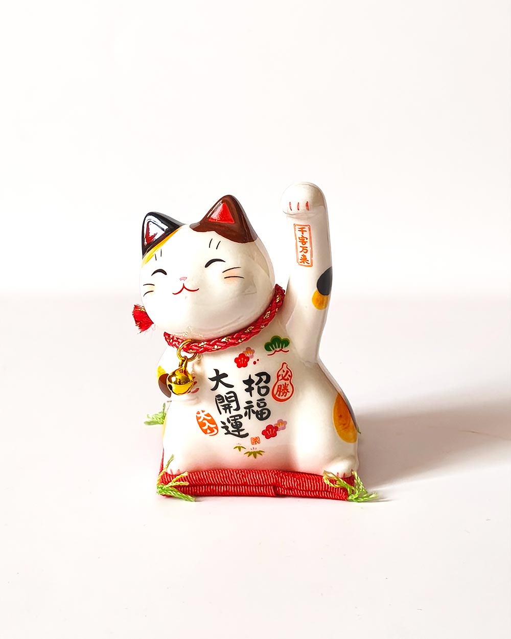 Small Japanese ceramic Maneki Neko lucky cat with raised paw, bell, colourful decorations and red cushion. Buy it on j-okini.com in Malta (Europe)