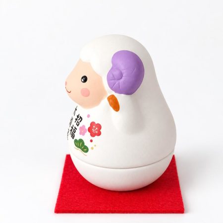Round white Japanese zodiac sheep figurine with purple horns, floral decoration and Japanese good luck lettering, displayed on a red mat.